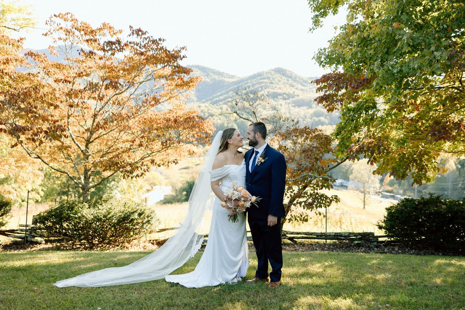 Guests gather outdoors for a scenic wedding ceremony in a lush garden setting under tall green trees.