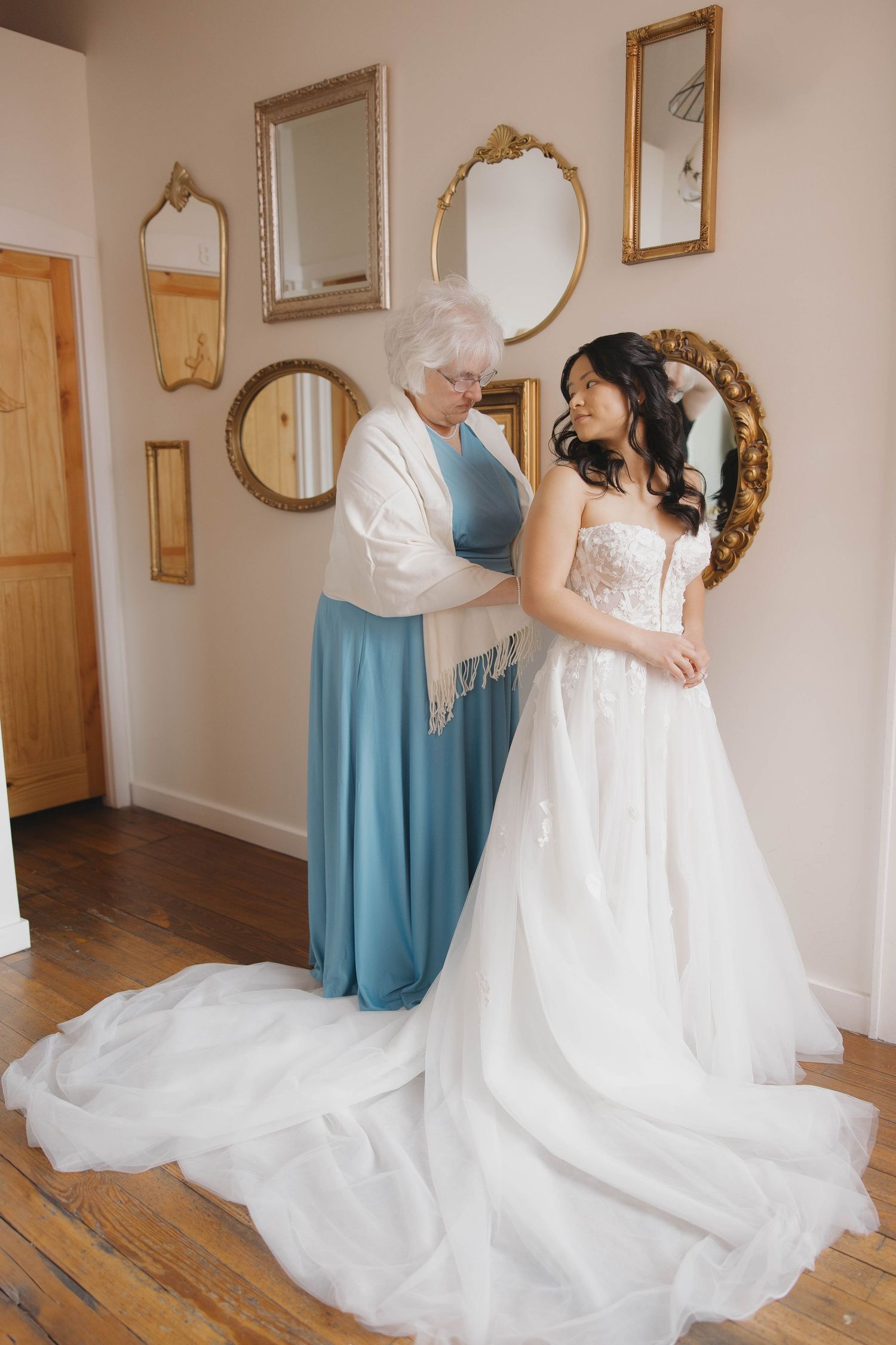 Two people stand together in an elegant dressing room with antique mirrors, one wearing a long white wedding gown.