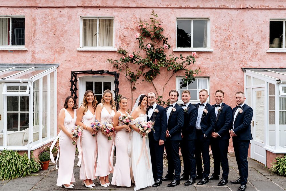 Bridal party in black tuxedos and blush dresses poses for formal photo against pink building exterior.