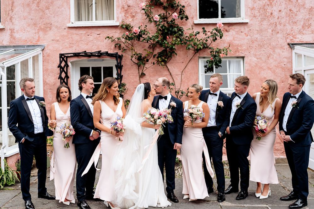 Wedding party poses together in formal attire against pink stucco wall with climbing vines.