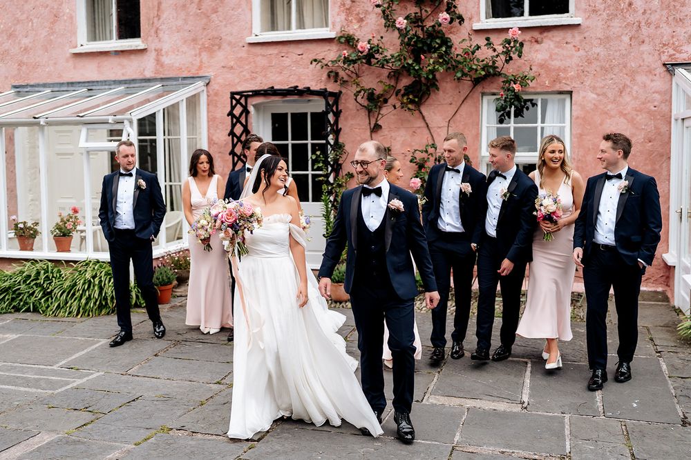Wedding party in formal attire walks together outside pink building during celebration.