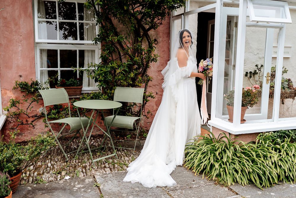 Bride in elegant white gown poses outside pink house with vintage patio furniture.