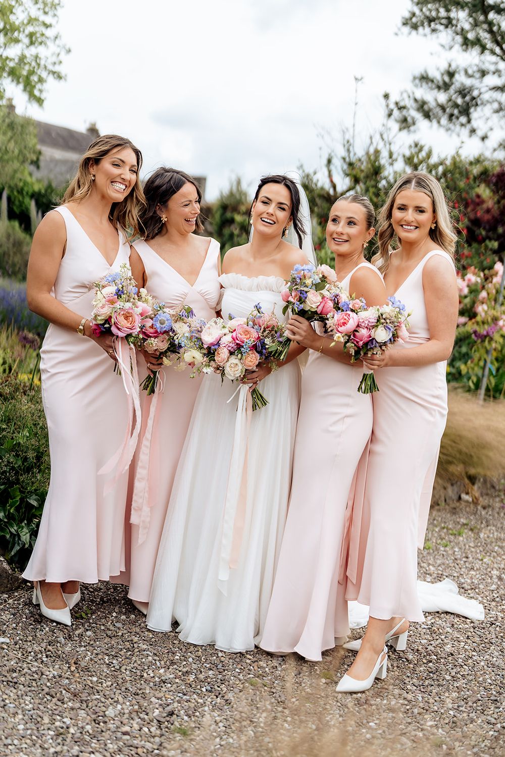 Bridesmaids in blush pink dresses pose with bride holding pink and white floral bouquets in garden setting.