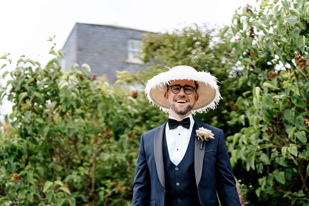 A stylish wedding attendant in a black tuxedo, white bow tie, and wide-brimmed straw hat stands among green foliage.