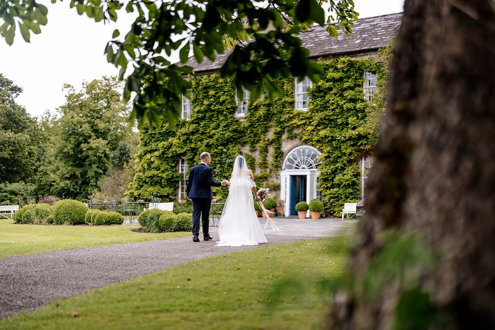 A couple walks along a landscaped garden path with trees and buildings in the background.