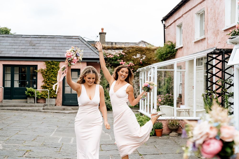 Three people in elegant white dresses joyfully celebrate outdoors at a stone patio venue with pink floral decor.