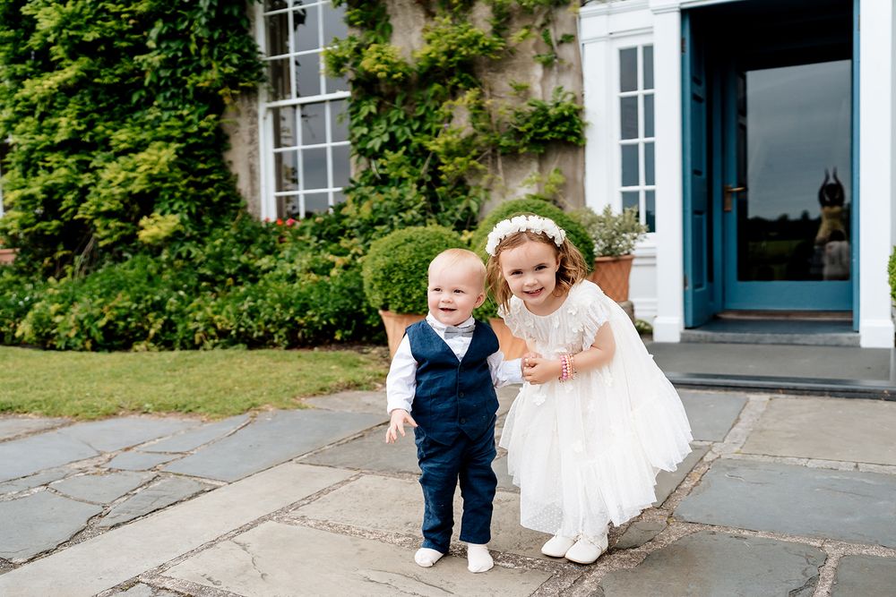 Little boy in navy suit walks alongside a woman in a white dress on an outdoors garden path.