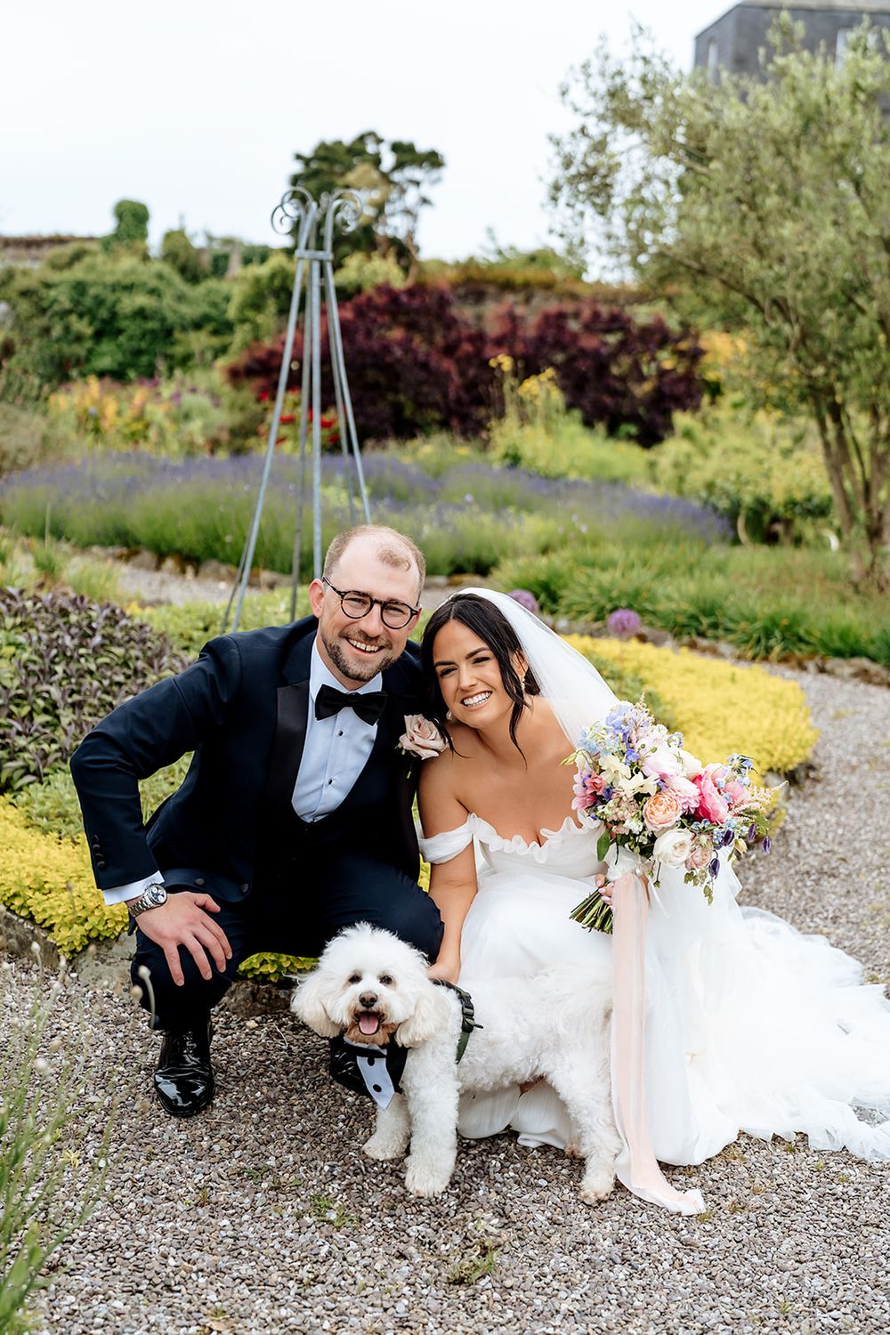 A bride and groom in formal wedding attire pose on a gravel path with their small white dog in a scenic garden setting.