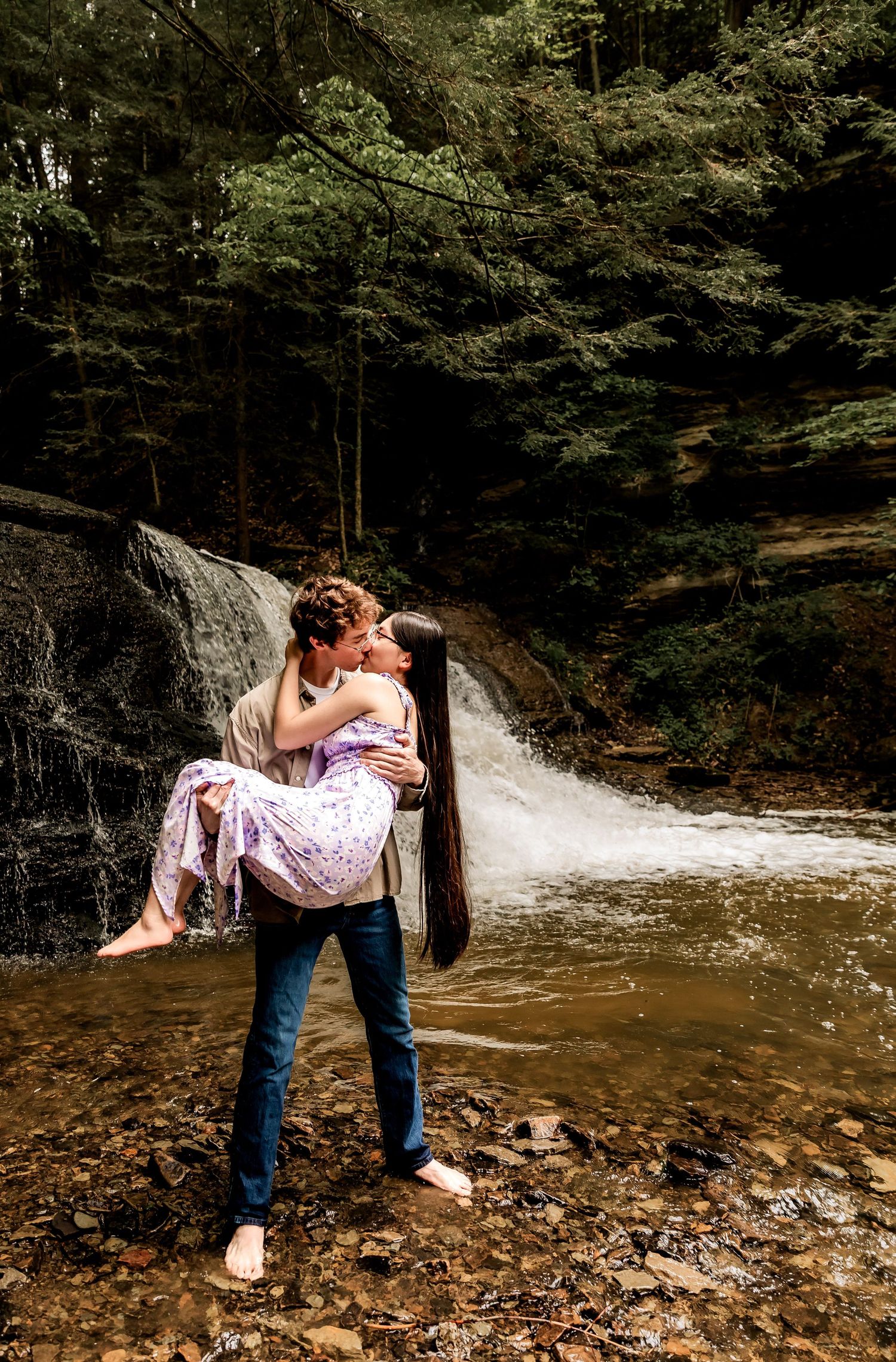 A couple embraces in front of a cascading waterfall in a lush forest setting.