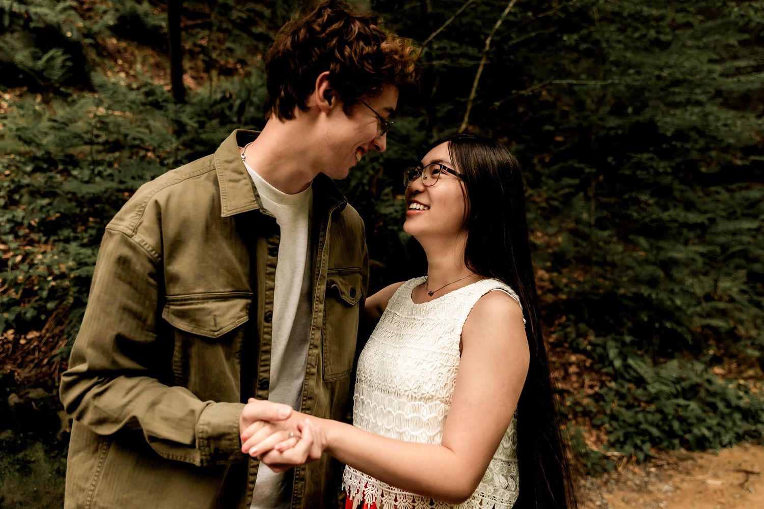 A couple shares a joyful moment together in the woods wearing tan jacket and white lace dress.