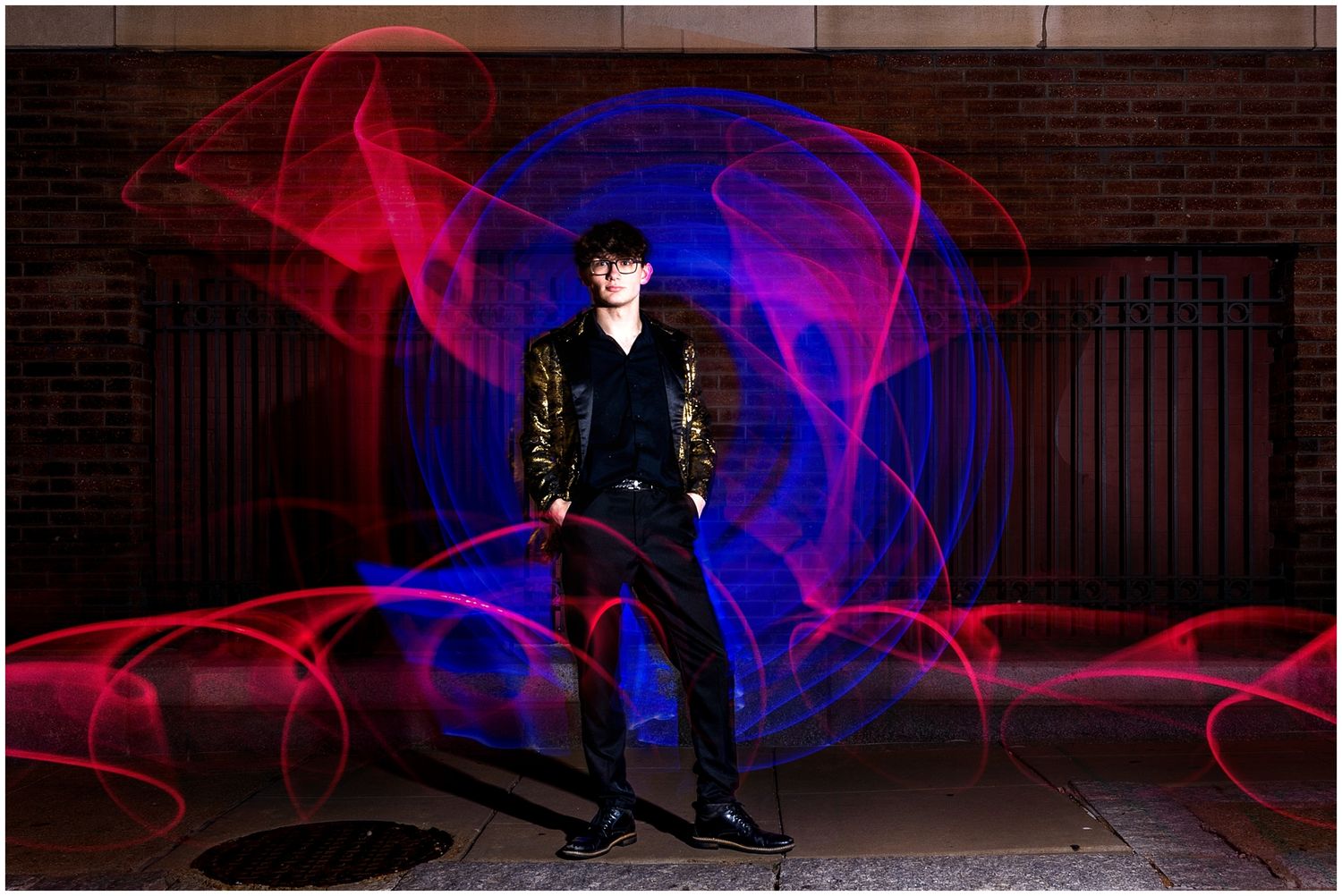 Dramatic light painting photograph with red and blue light trails swirling around a figure in black against brick wall.