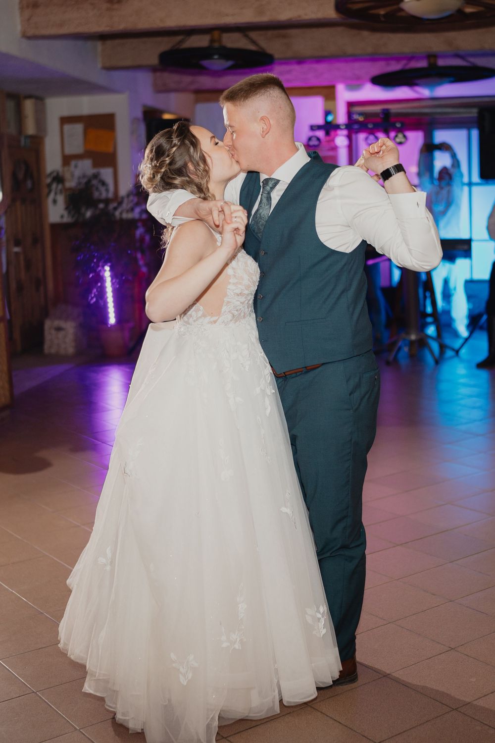 A couple in formal wedding attire shares their first dance in a venue with purple lighting and tiled floor.