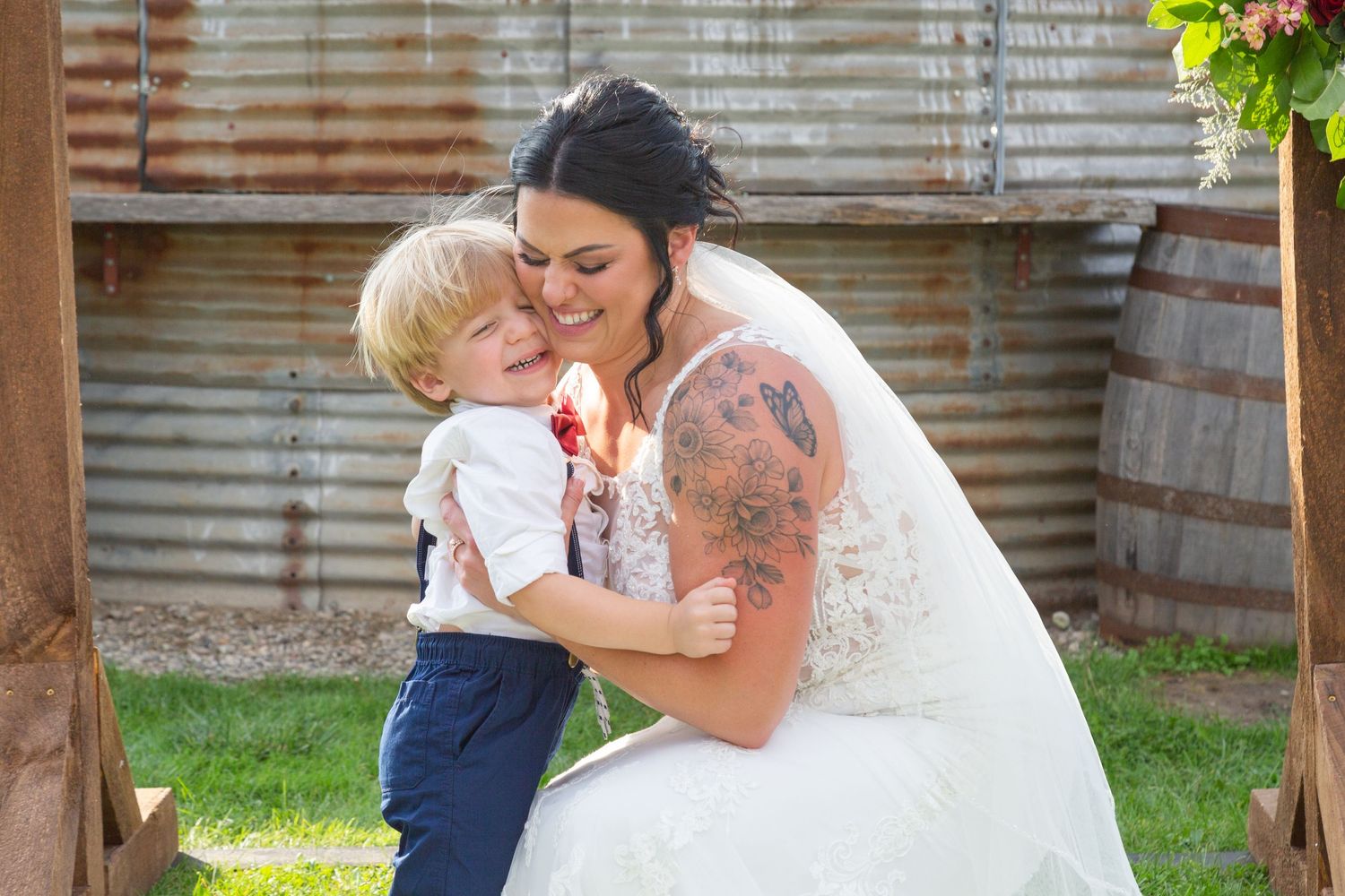 A bride in a white wedding dress with tattoos shares a tender moment with a young child in blue outfit outdoors.
