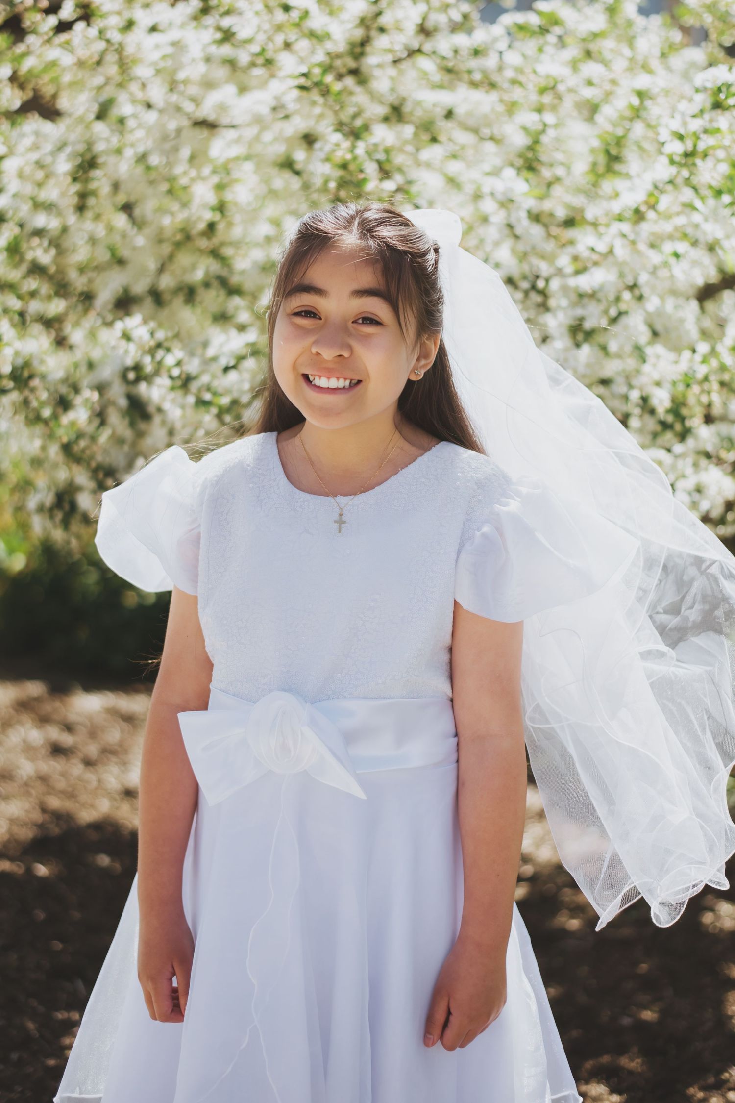 A girl wearing a white dress with a veil stands in front of blooming spring flowers on a sunny day for her confirmation and first communion.