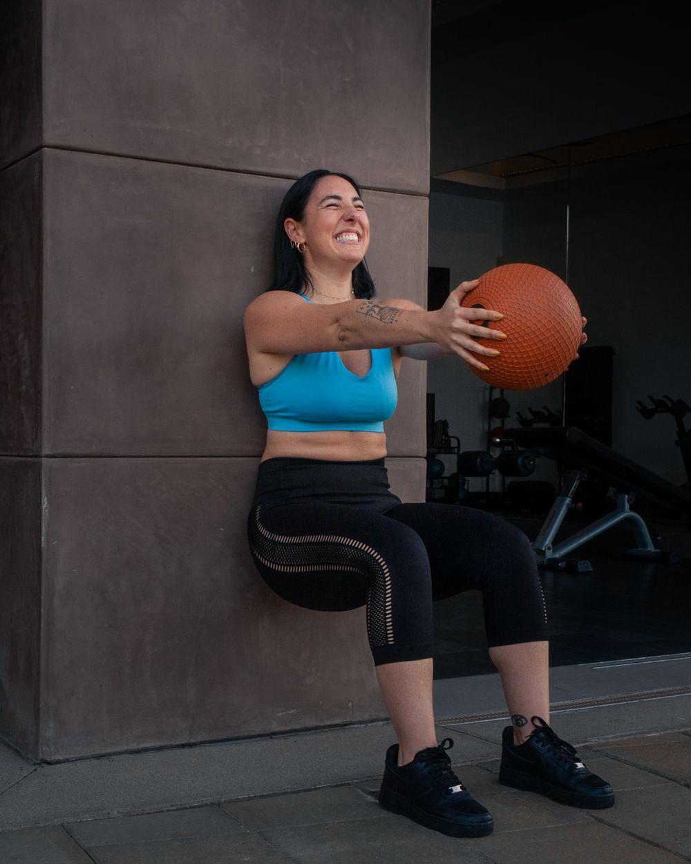 An athlete in blue workout attire smiles while holding a medicine ball during a wall-sit exercise. Brand photography, San Diego, California