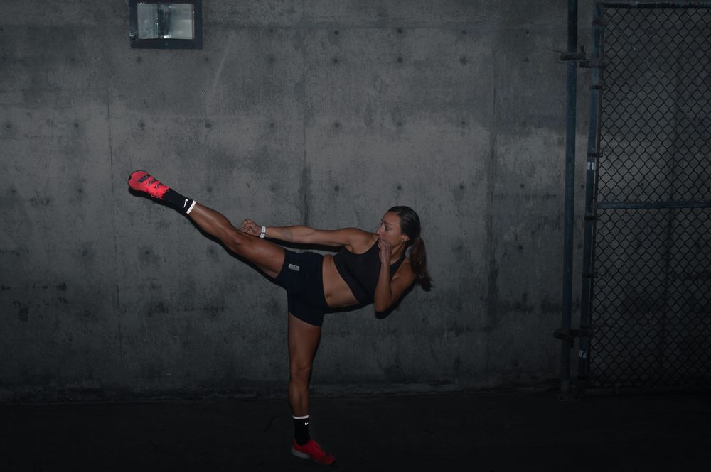 An athlete performs a high kick in dark workout gear against a concrete wall. Brand photography, San Diego, California