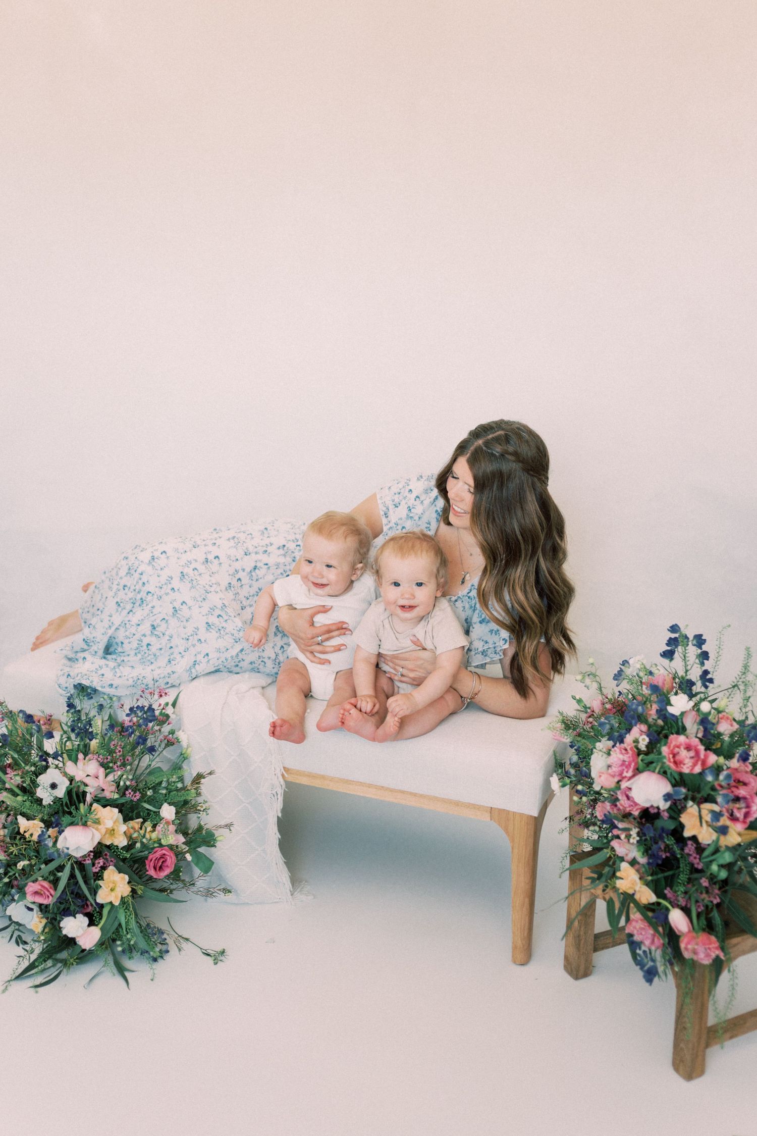 Family portrait on white settee surrounded by pink and purple floral arrangements on a pale backdrop.