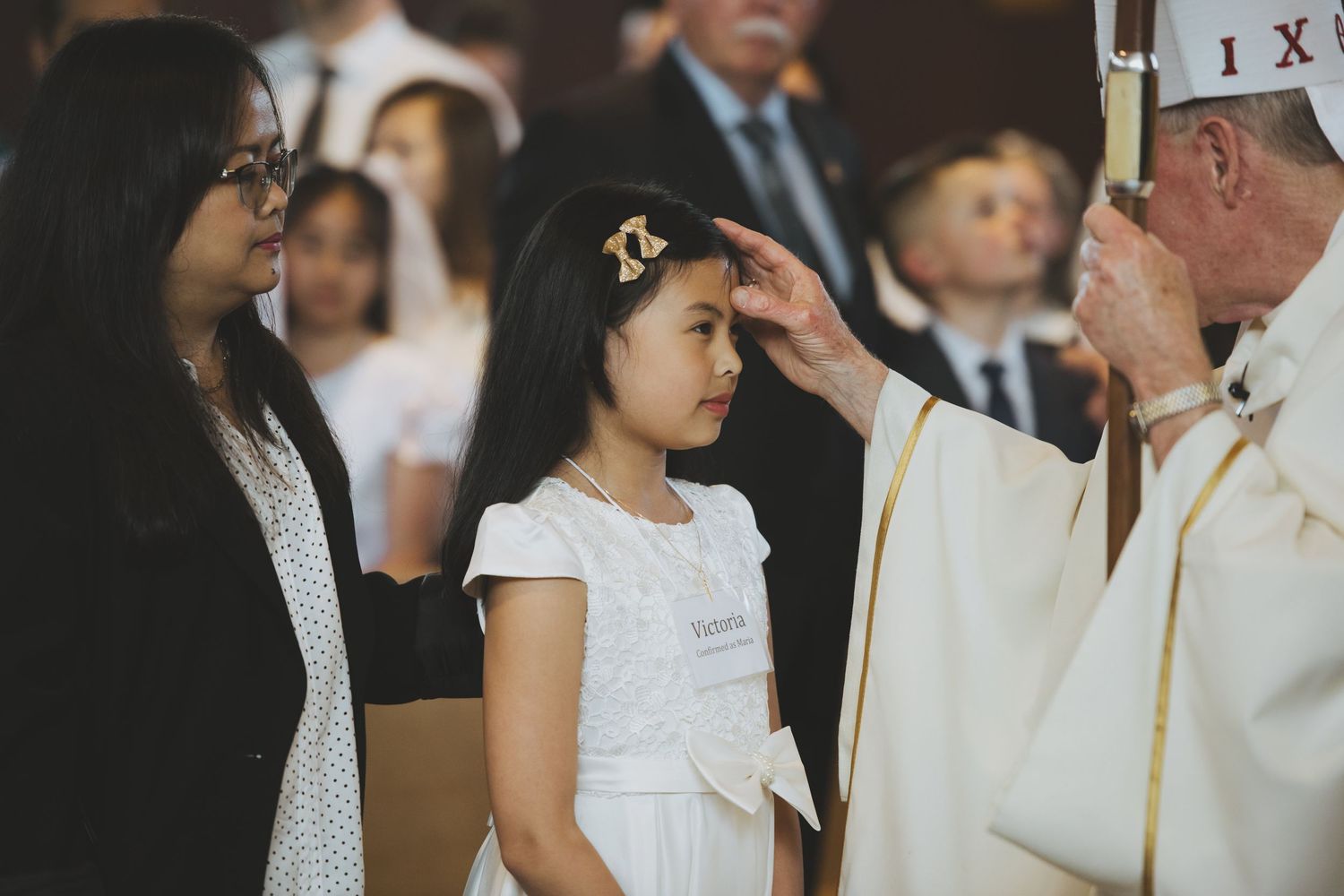 A young person in a dark suit participates in a religious ceremony as clergy performs a blessing.