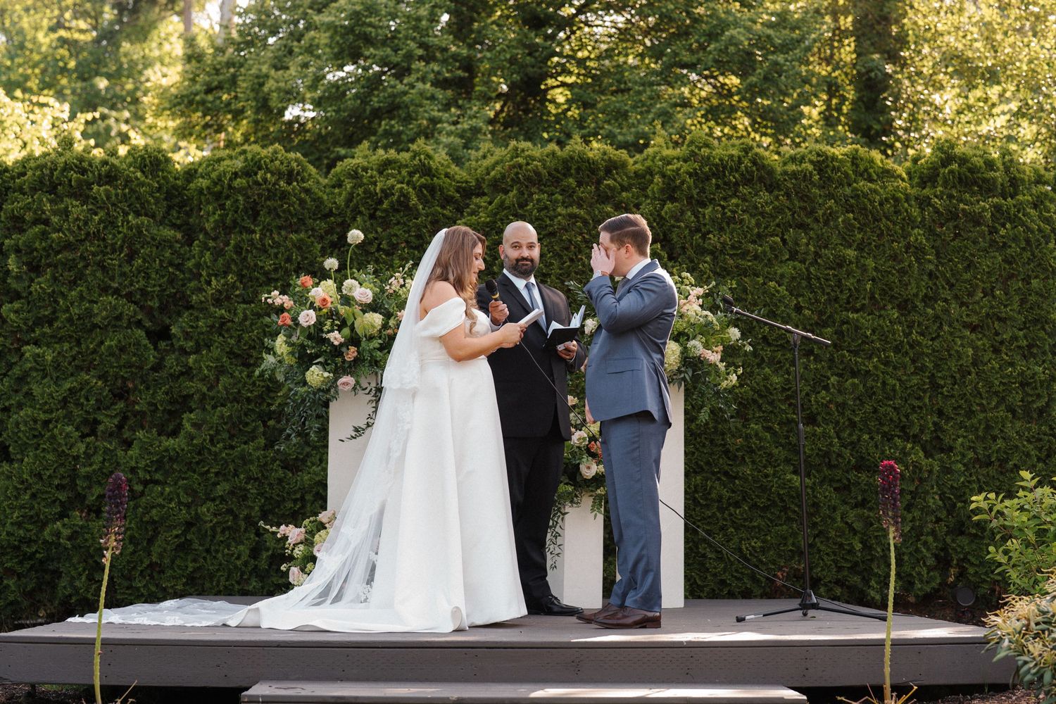 Outdoor wedding ceremony takes place on elevated platform with floral arrangements and hedge backdrop in garden setting.