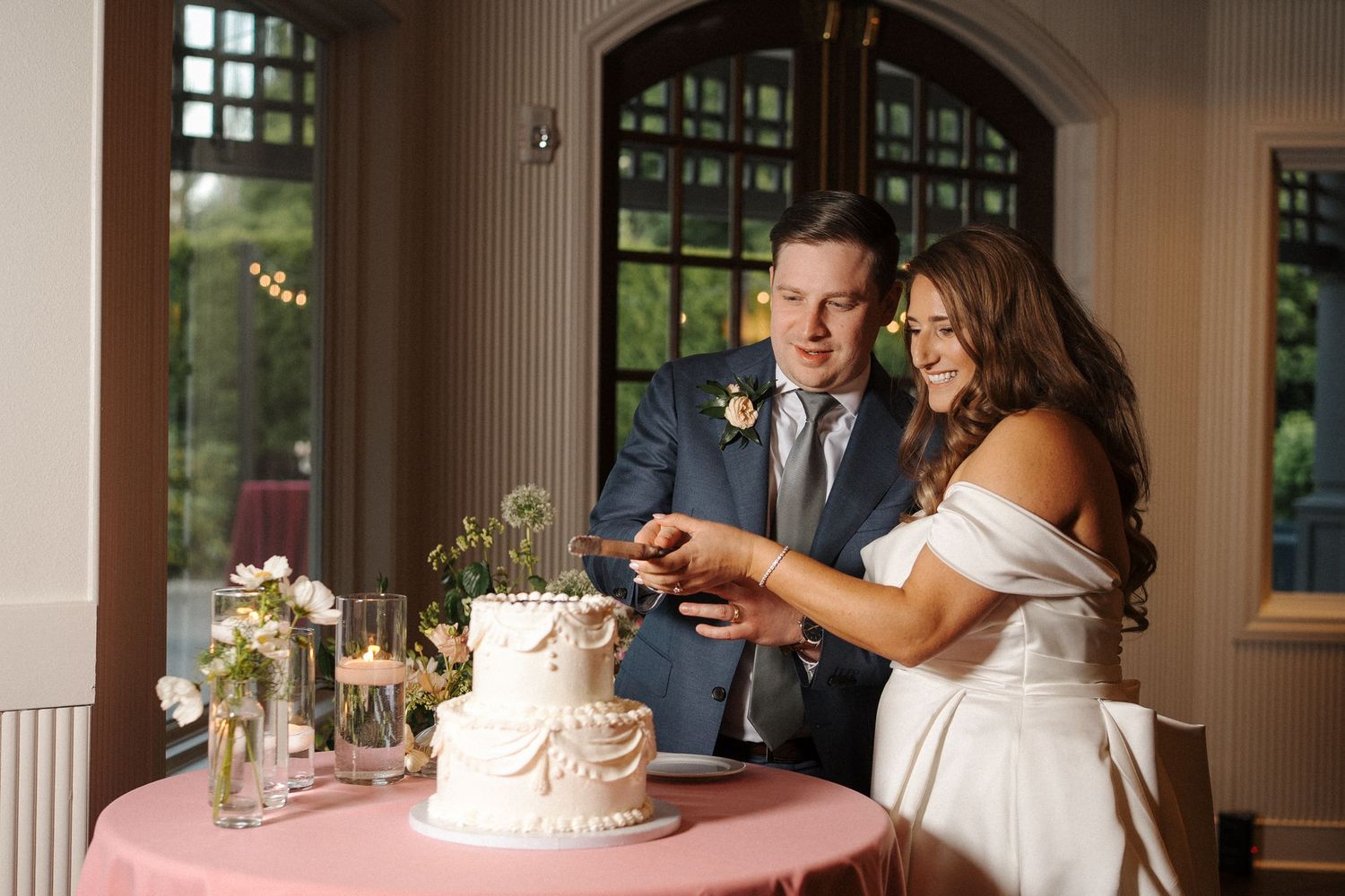A couple cuts into a white textured wedding cake together at an elegant indoor venue with arched windows.