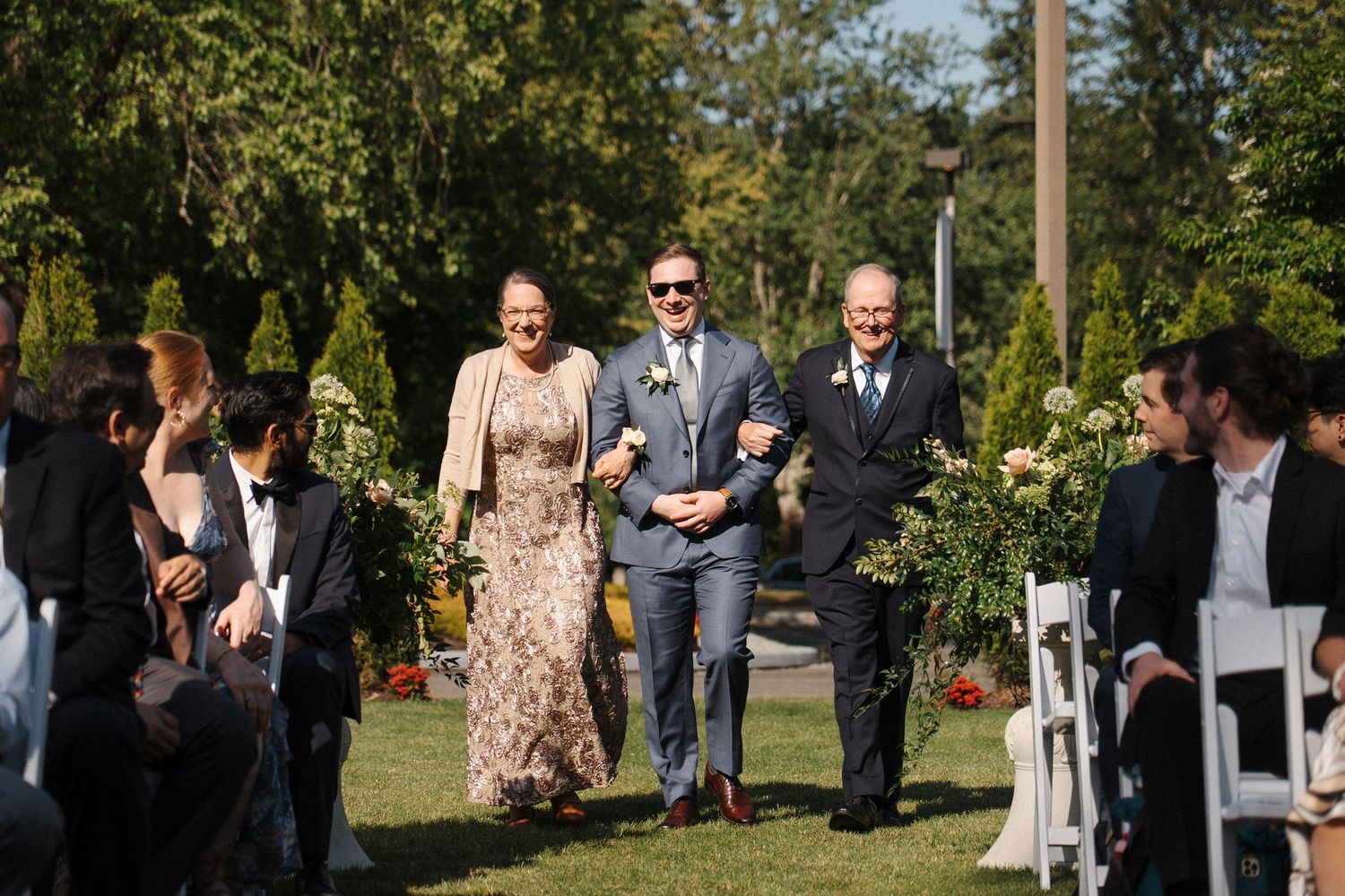 Wedding guests enjoy an outdoor ceremony seating area on a sunny day with green trees in the background.