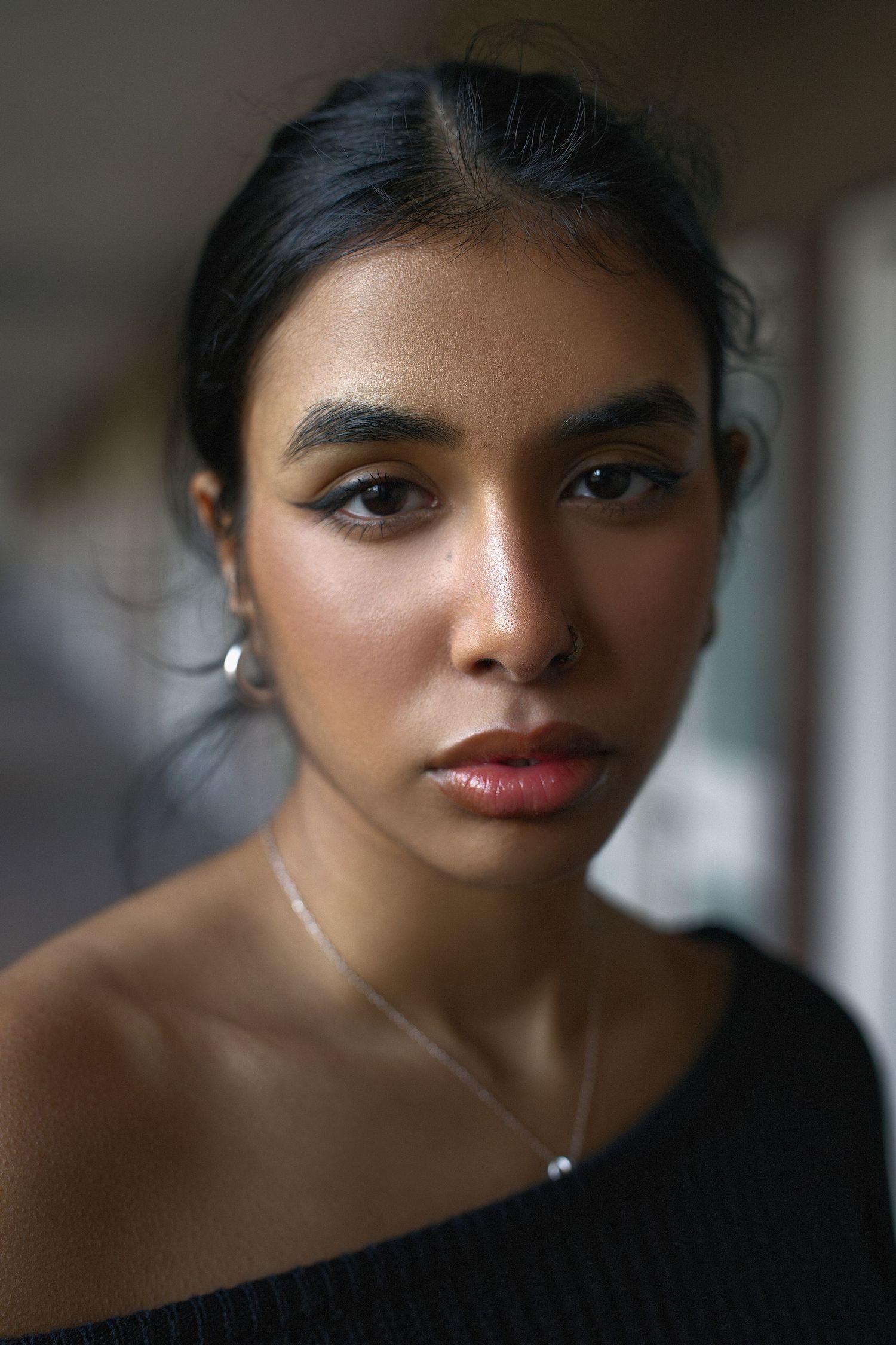 Portrait photo showing beautiful South Asian woman in black off-shoulder top with delicate necklace against blurred background.