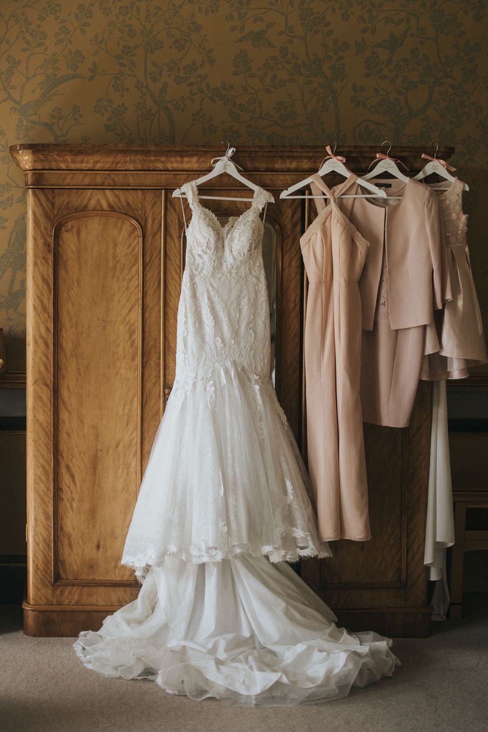 A wedding dress and blush bridesmaid dresses hanging elegantly in a rustic room against an antique wooden cabinet at Rise Hall.