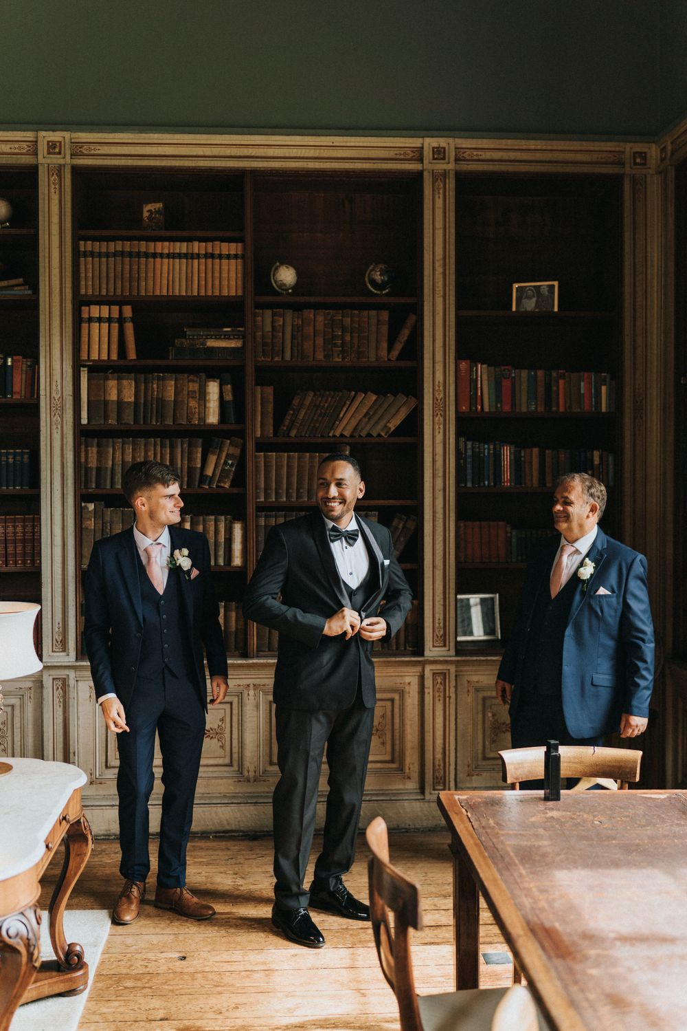 Three men in formal black tuxedos share a lighthearted moment in an elegant library setting with wooden bookshelves at Rise Hall.