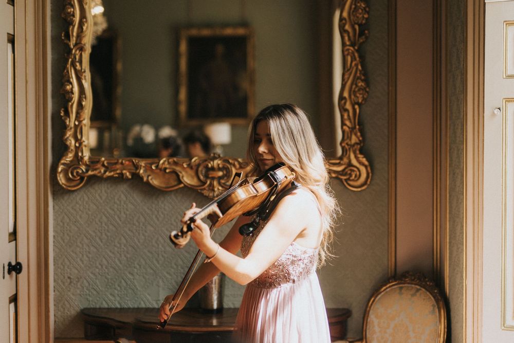 A person in a white dress plays violin in a vintage room with an ornate gold mirror at Rise Hall.