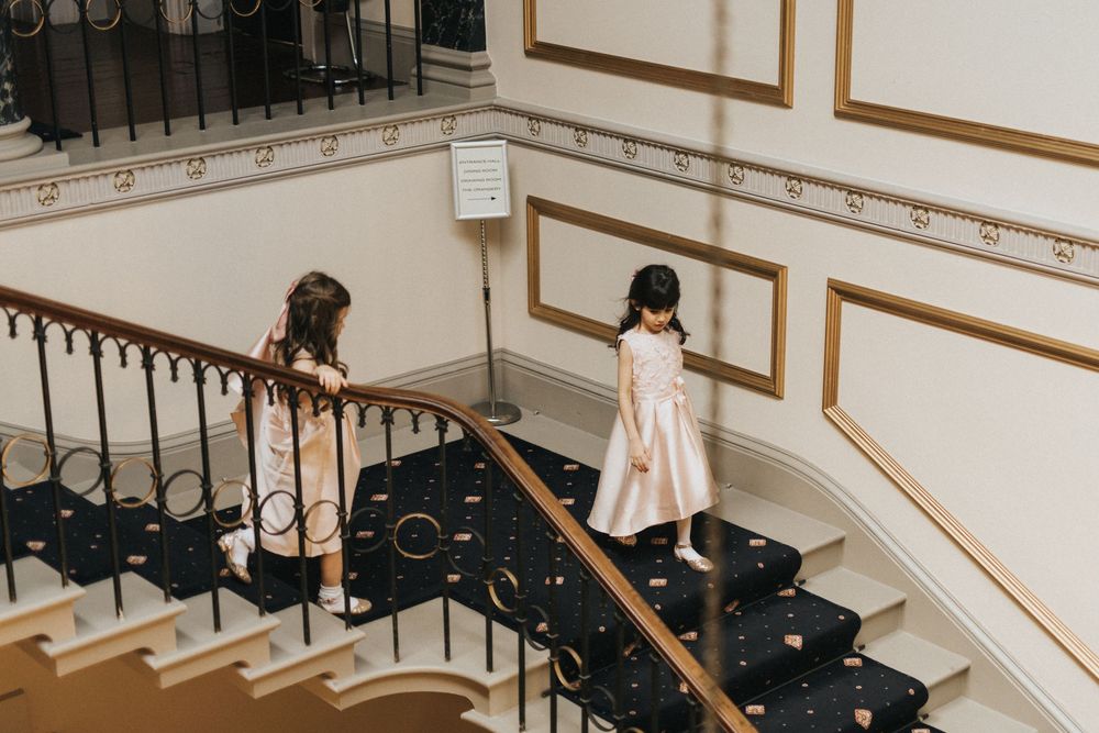 Two wedding guests walk down an elegant spiral staircase with ornate black iron railings and white walls at Rise Hall.