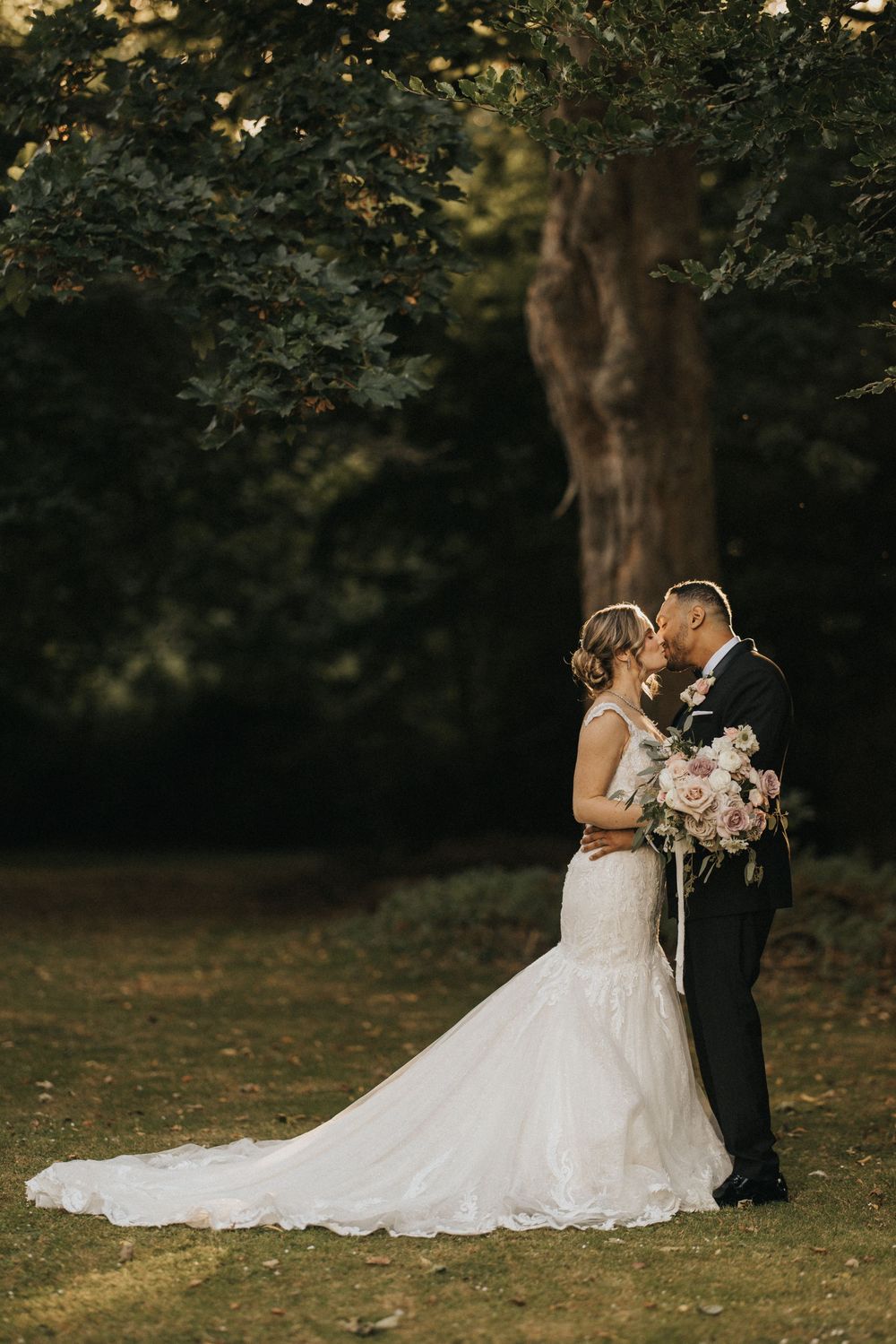 A romantic wedding couple embraces outdoors under trees in a natural garden setting at dusk at Rise Hall.