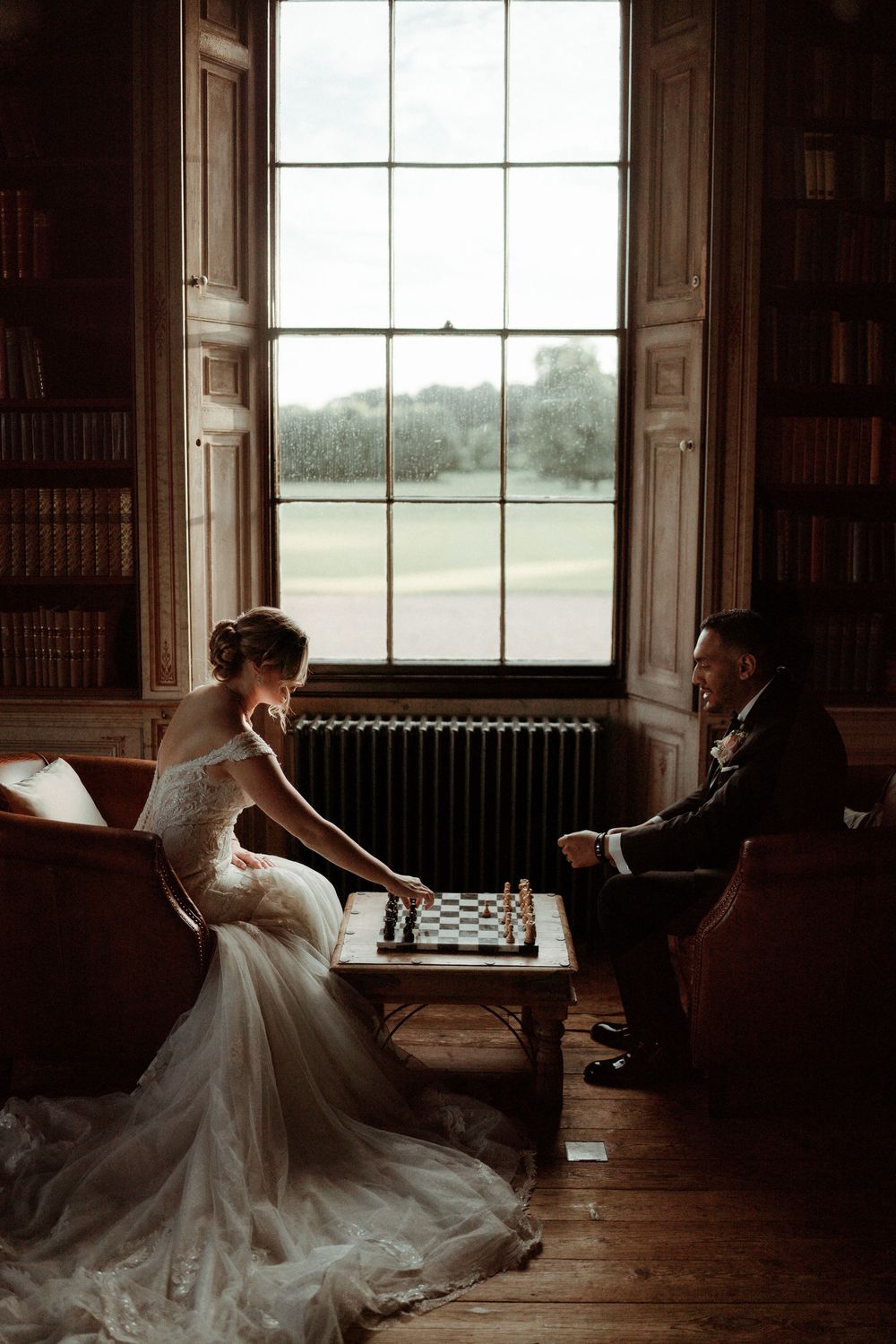 A couple in wedding attire play chess by a tall window in a vintage library setting at Rise Hall. Taken by Grimsby photographers.