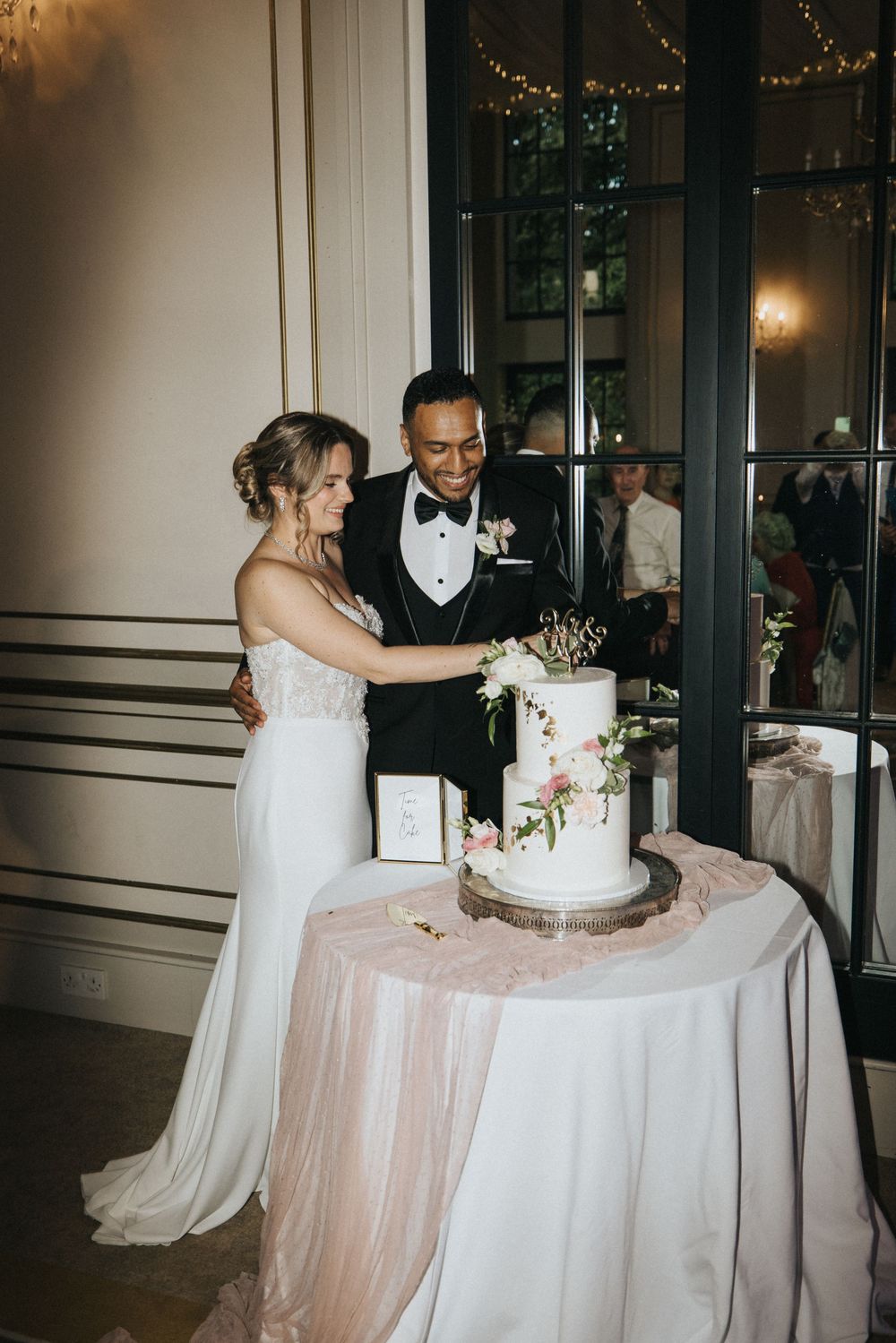 A couple cuts their elegant white wedding cake adorned with floral decorations on a pink and white draped table at Rise Hall.