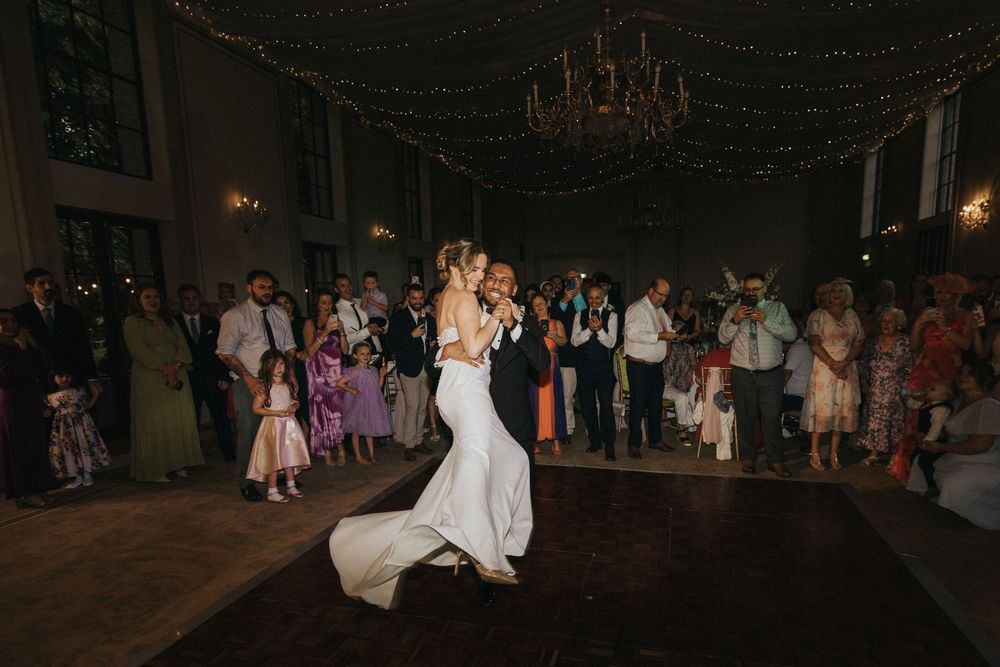 A couple shares their first dance at an evening wedding reception at Rise Hall beneath a chandelier while guests watch from the sidelines.