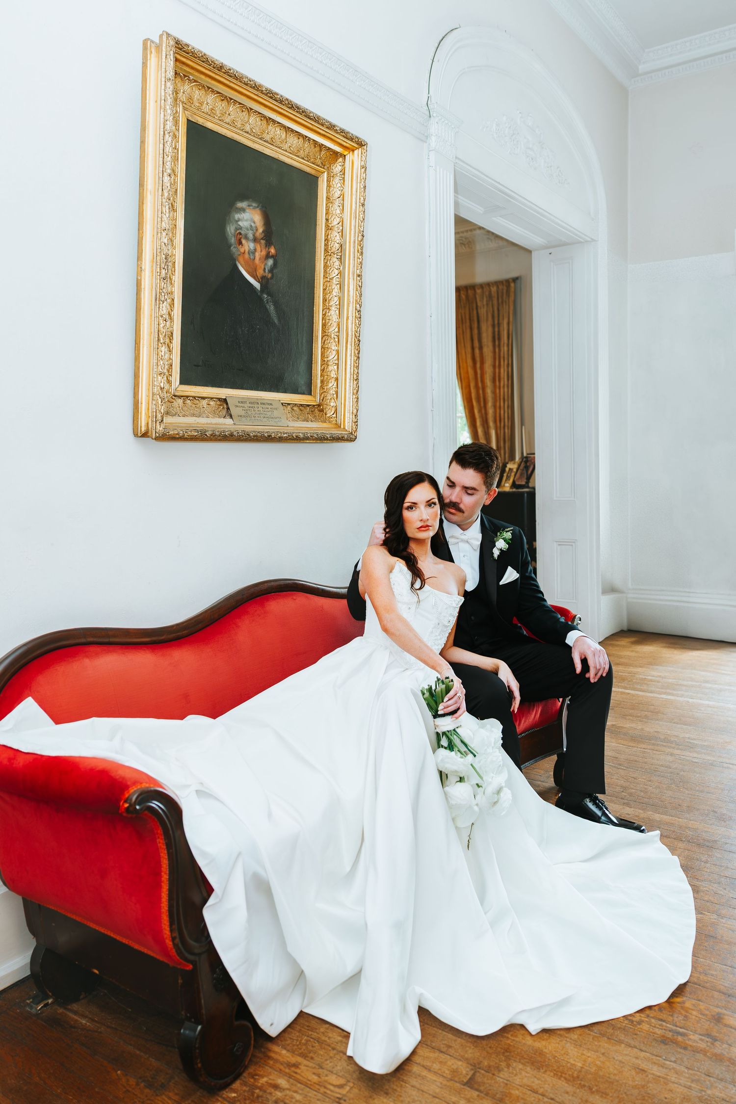 A series of romantic wedding photos with a bride and groom posing on an elegant red chaise lounge in a bright gallery room.