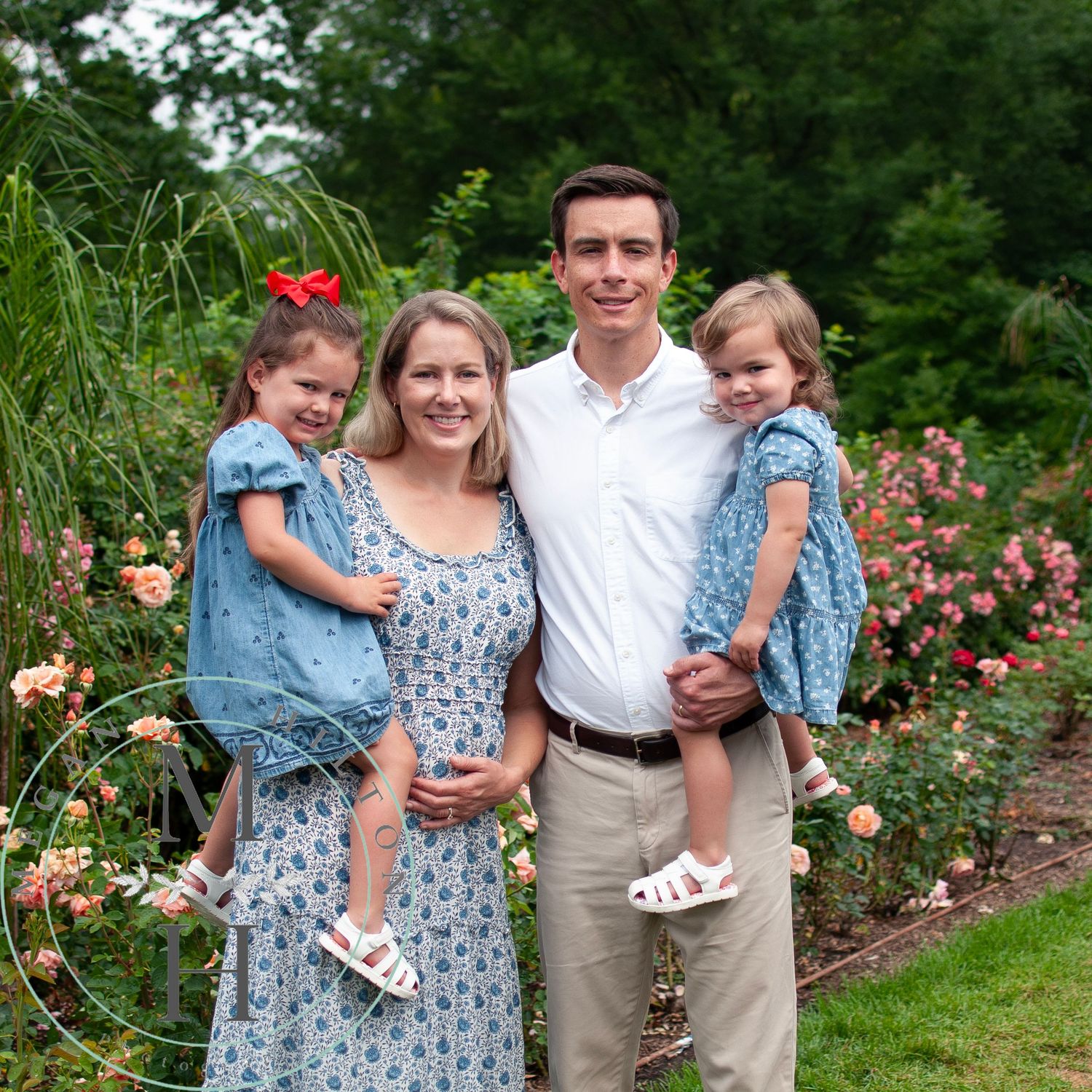 A family of four poses in a garden with blooming pink roses and flowering plants in the background of their outdoor portrait.
