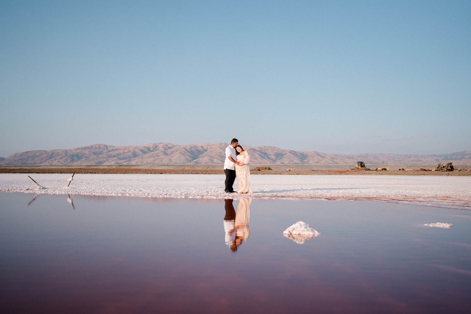 Two people stand on salt flats with mountains in background reflected in still water at sunset.
