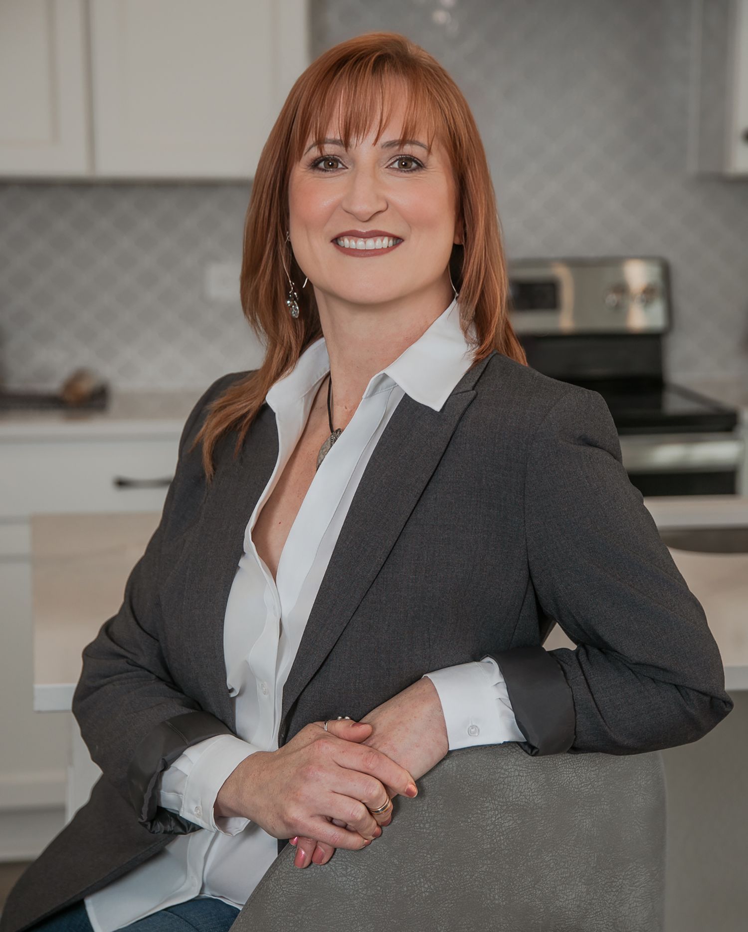 Professional woman in gray suit jacket smiling and posing in modern kitchen with stainless steel appliances.