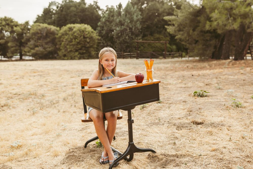 Student sitting at vintage school desk in outdoor setting with open book and drink on sunlit summer day.