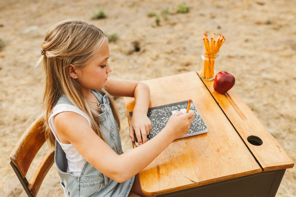 Student writing at wooden desk with orange pencil storage compartment and sun-filled background.