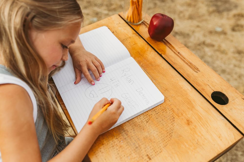 Student writing in notebook while sitting at wooden desk with red apple and pencil holder.