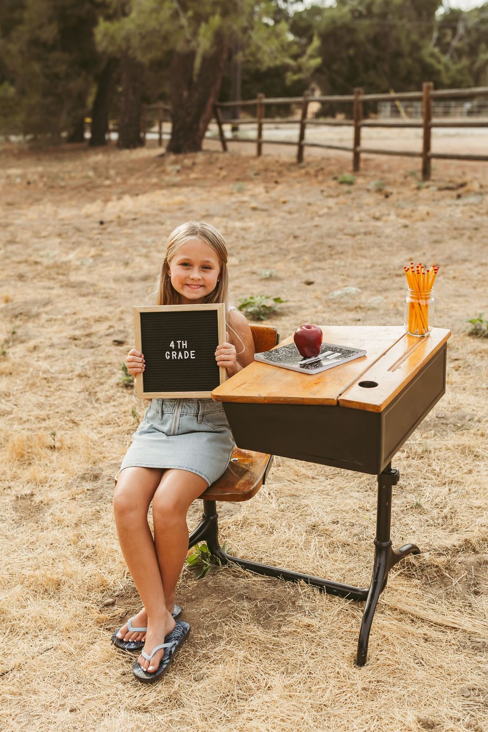 A student sits at a vintage school desk outdoors with a chalkboard sign and school supplies on a warm fall day.