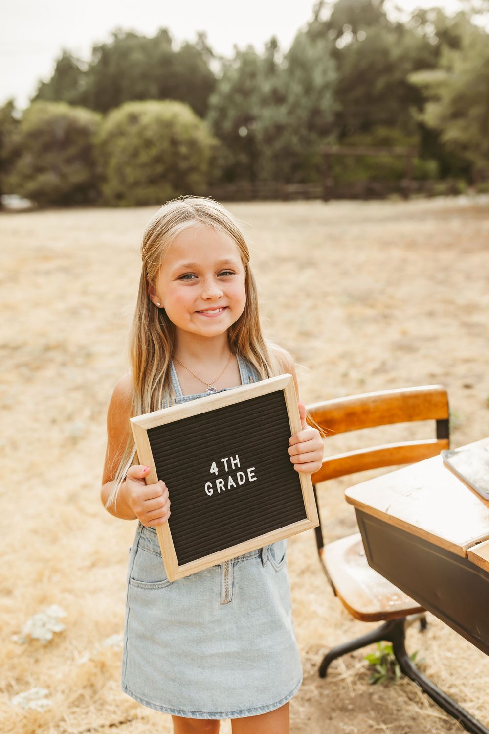 A letter board with '4th grade' text is held up outdoors against a natural background on a sunny day.