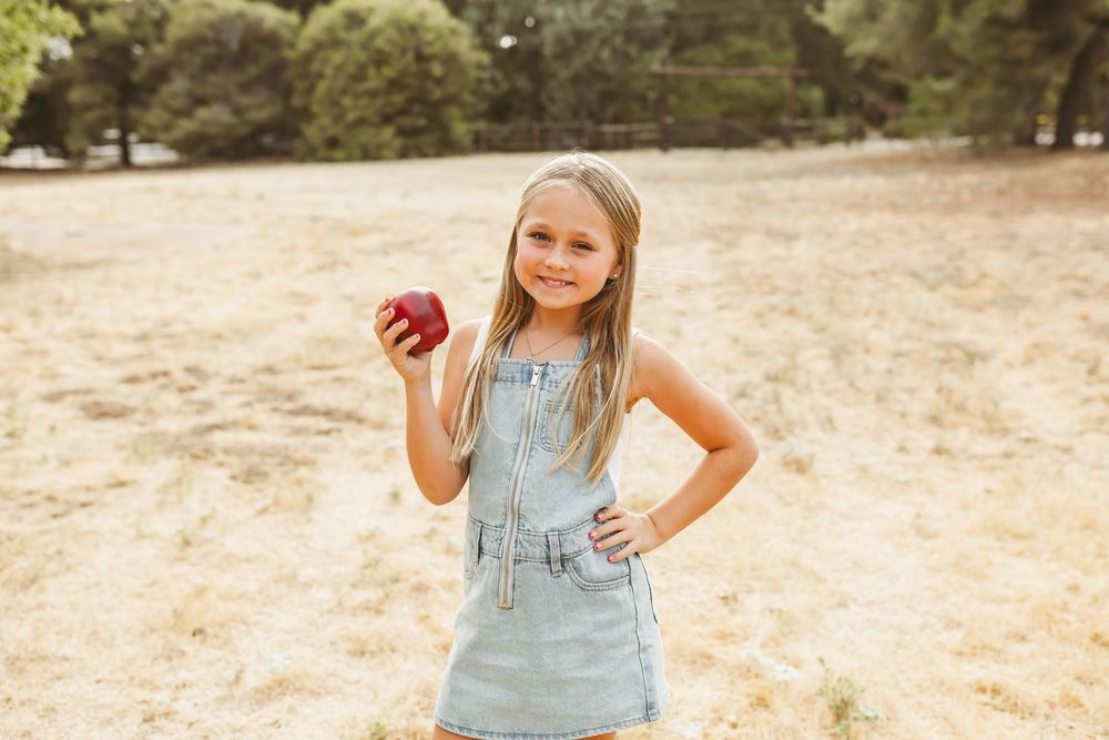 A child poses confidently in a denim overall dress while holding a bright red apple in an outdoor setting.