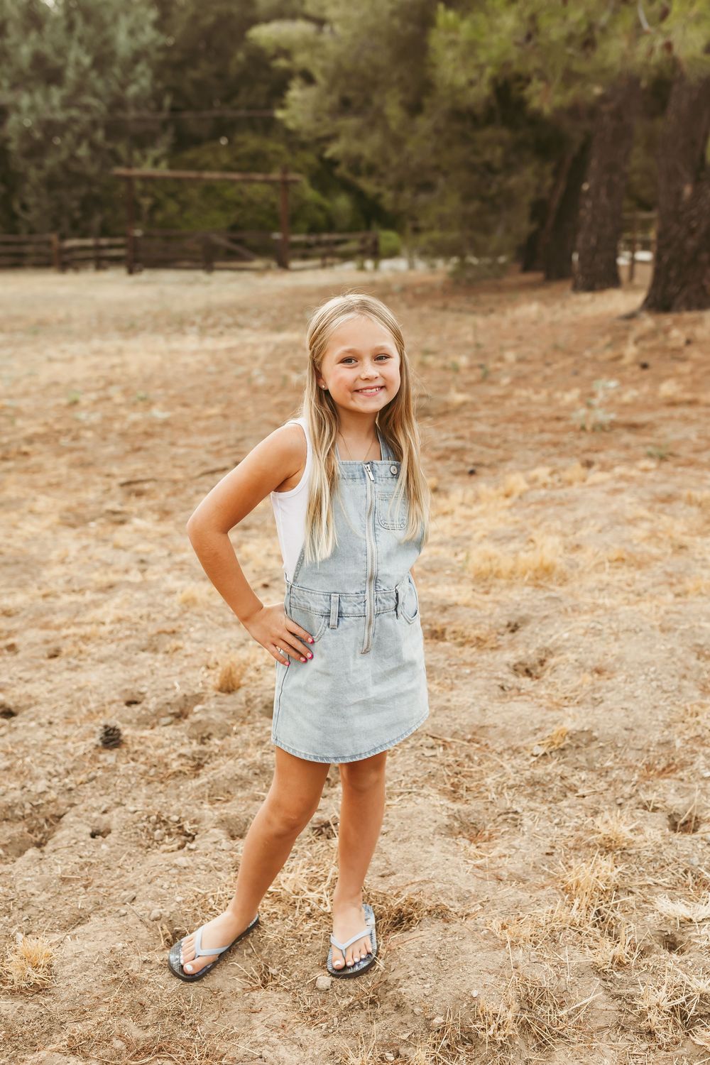 Young child in denim dress and sandals poses outdoors in casual summer style against a natural backdrop.