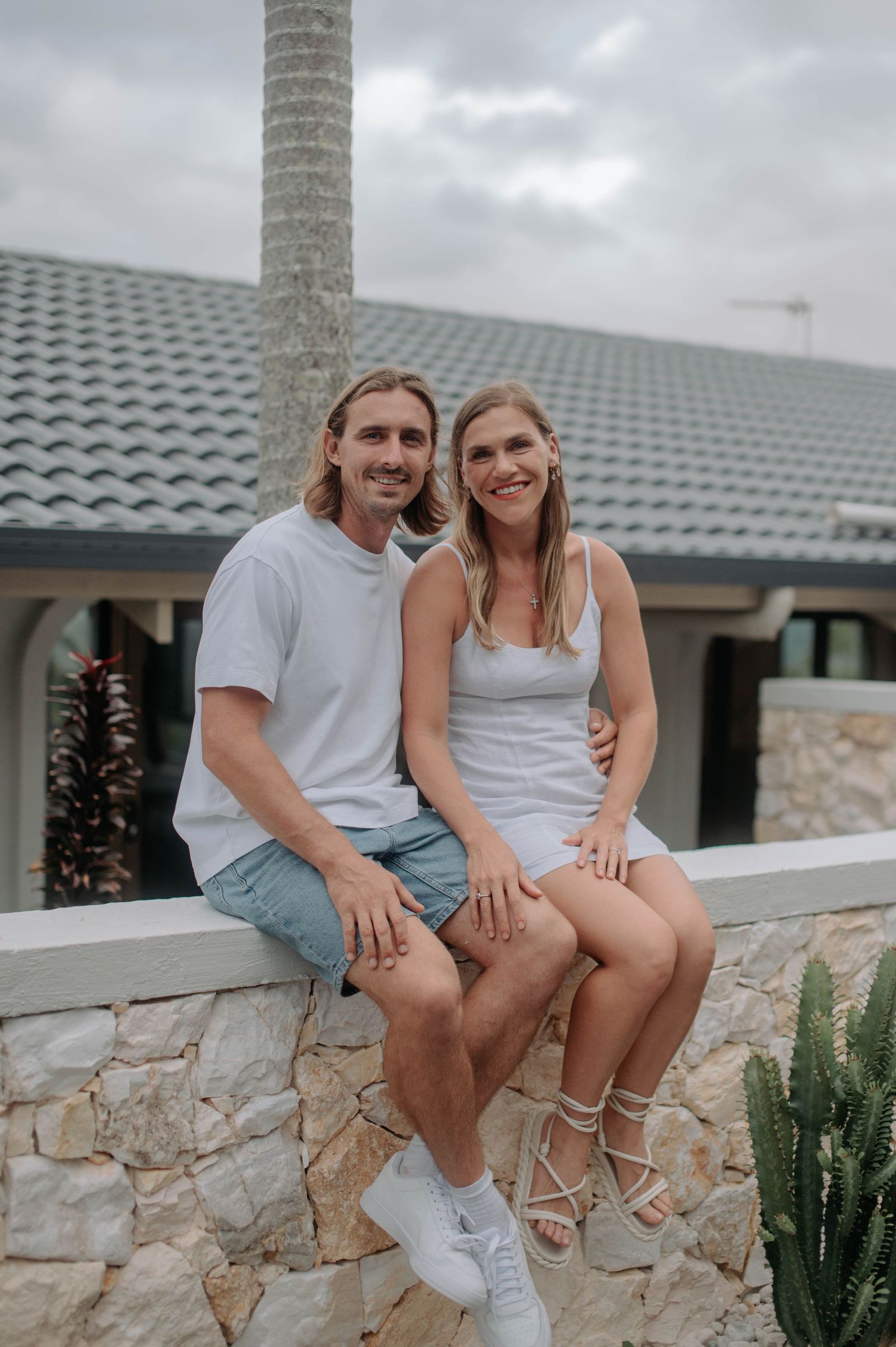 Two people in casual attire sit together on a stone wall outside a house with a tiled roof on a cloudy day.
