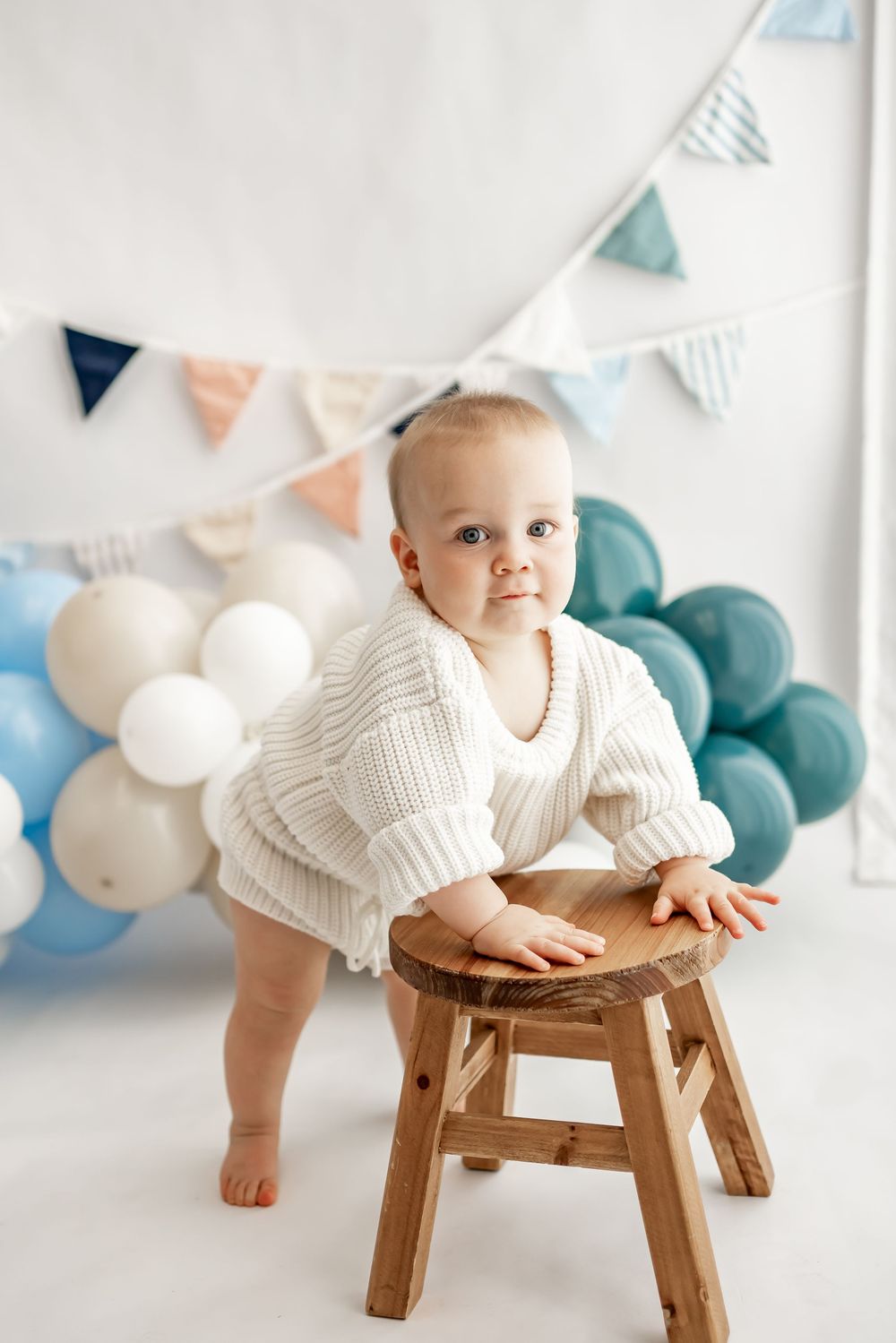 Festive first birthday party setup with blue and white balloons, bunting flags, and a small wooden stool against white backdrop.
