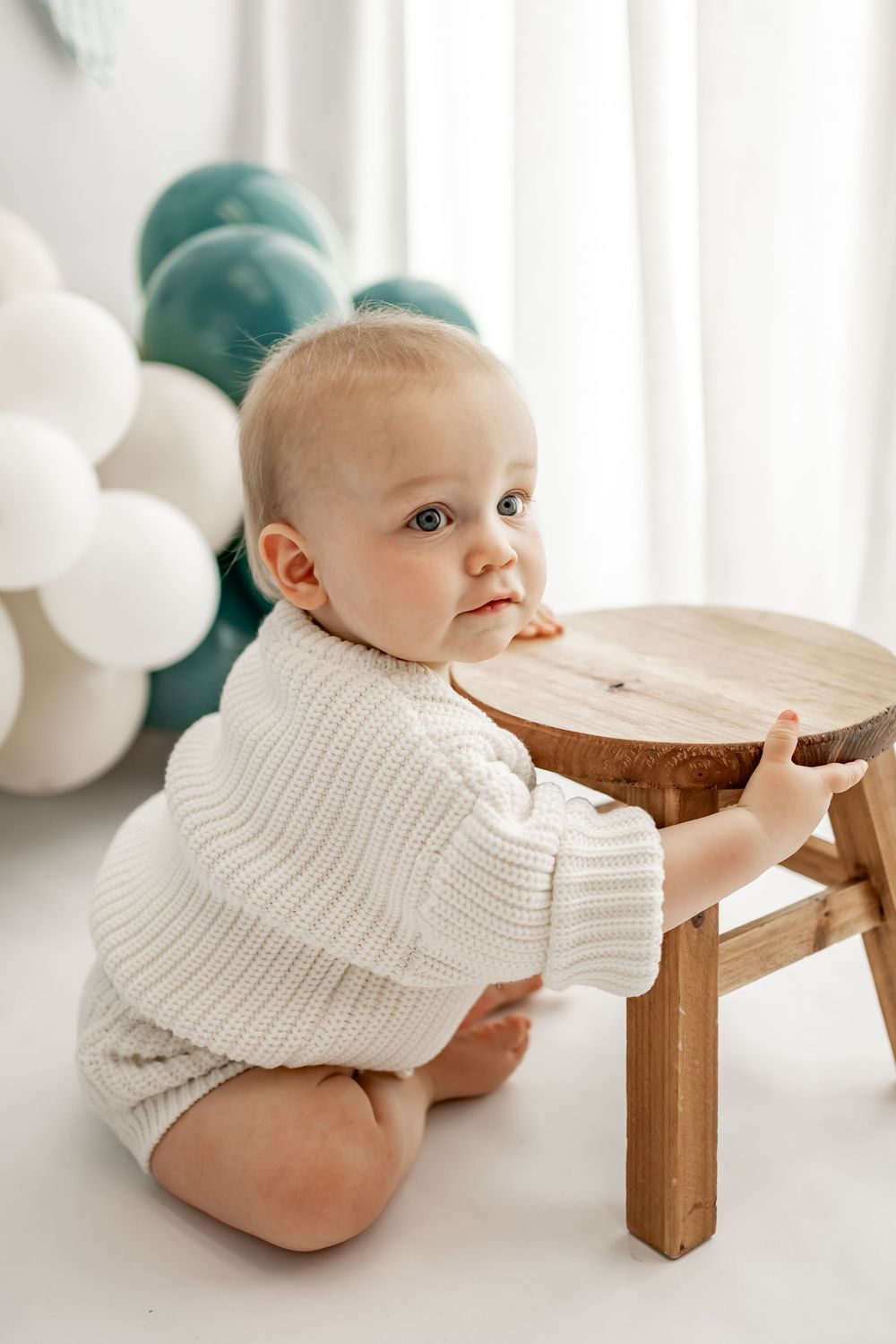 Baby in white knit sweater sits near wooden stool with turquoise and white balloons in background.