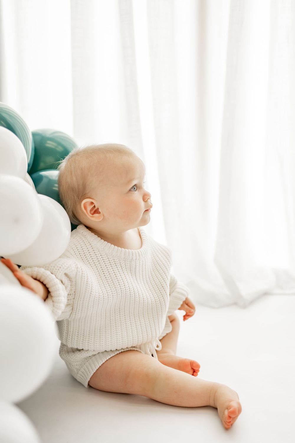 Baby in white knit sweater sits beside balloons against bright window with soft natural lighting.
