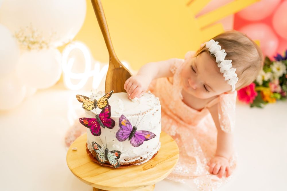 Elegant first birthday cake decorated with butterflies and pink accents next to golden balloons and floral arrangements.